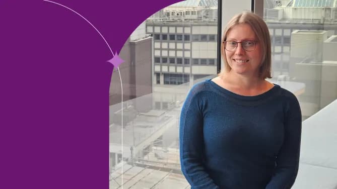 Debra is standing in front of a window with a city backdrop wearing a royal blue sweater and glasses. 