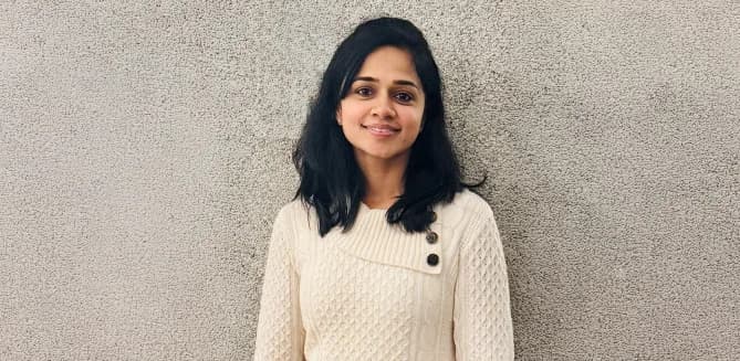Kavitha is standing in front of a tan wall, in a white sweater, smiling. 