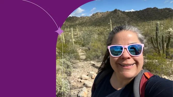 Stacy is standing in the desert wearing pink reflective sunglasses. Sage green bushes and tall cacti cover the mountains behind her.